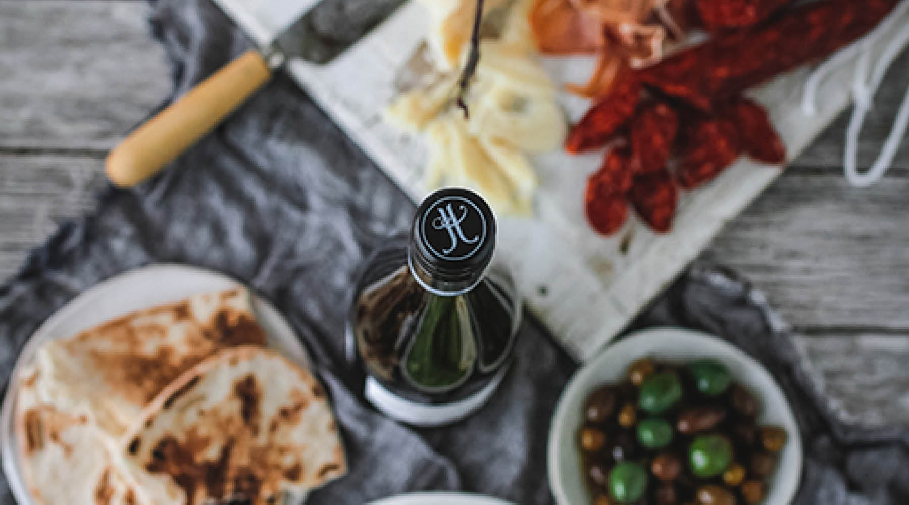 A top-down view of a bottle of Jules Taylor Gruner Veltliner wtih the Jules Taylor monogram clearly in view, with snacks including pita breads, green olives, and a charcuterie board on a gray cloth blurred in the background on a weathered wooden table.