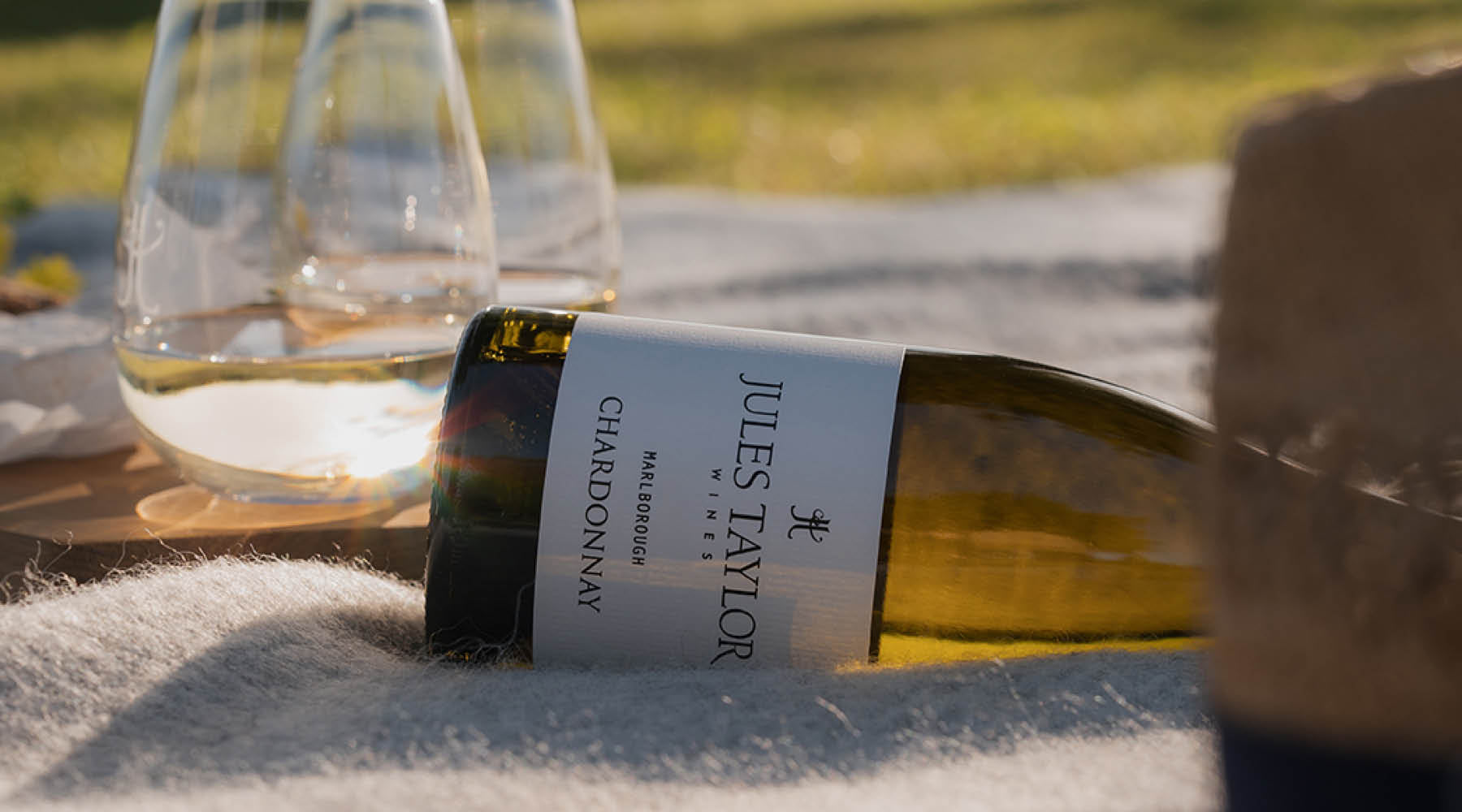 A bottle of Jules Taylor Chardonnay wine lies on a white blanket outdoors, with two wine glasses and a blurred outdoor background.