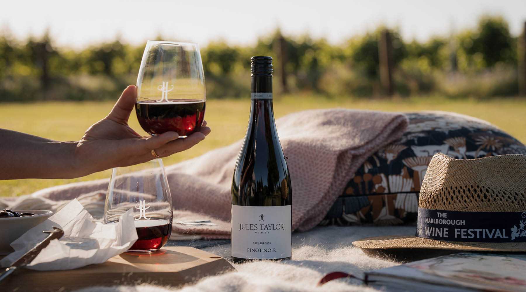 A bottle of Jules Taylor Pinot Noir wine and two wine glasses on a picnic blanket outdoors, with a hand holding one glass. A straw hat and a blanket are also visible, with a blurred vineyard in the background.