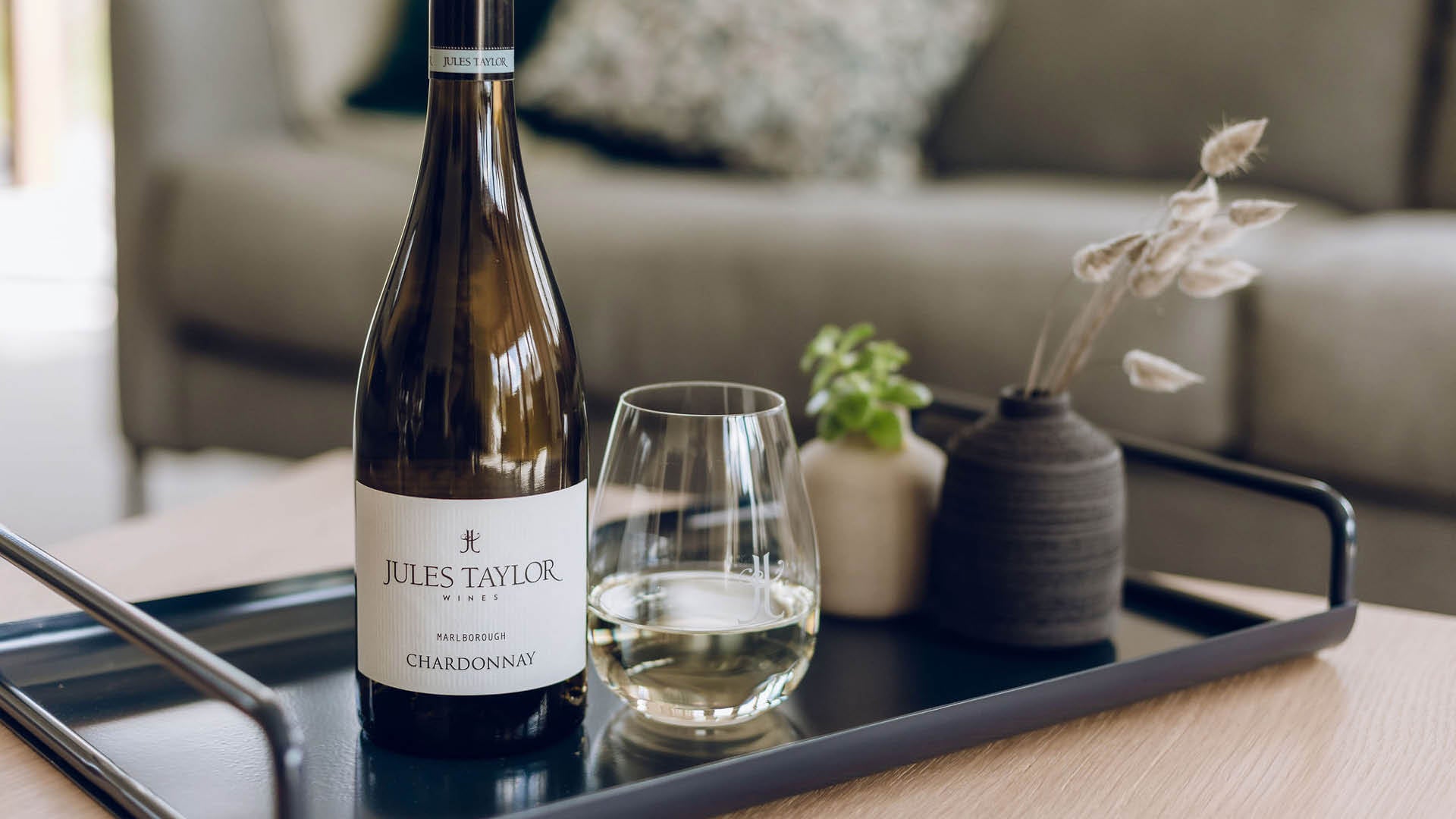 A bottle of Jules Taylor Chardonnay wine and a glass of white wine on a dark tray in a cozy living room setting.
