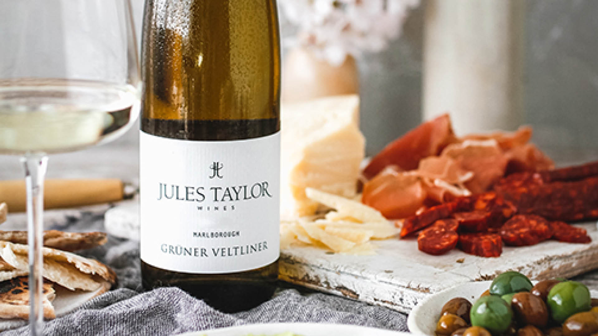 A bottle of Jules Taylor Gruner Veltliner white wine stands on a table beside a glass of white wine, with cheese, cured meats, and olives in a styled dining setting.