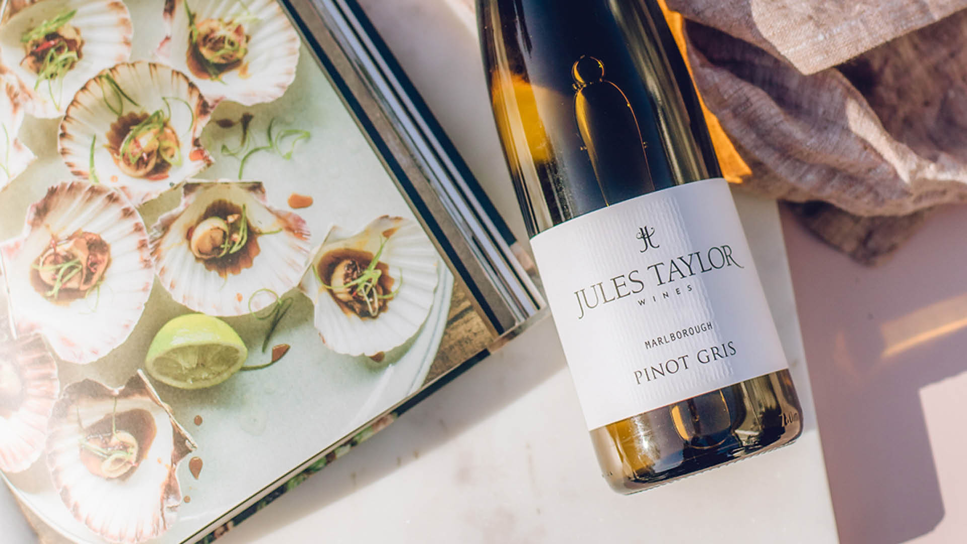A bottle of Jules Taylor Pinot Gris white wine lies diagonally on a light marble surface next to a printed cookbook featuring scallop dishes and a beige textured cloth.