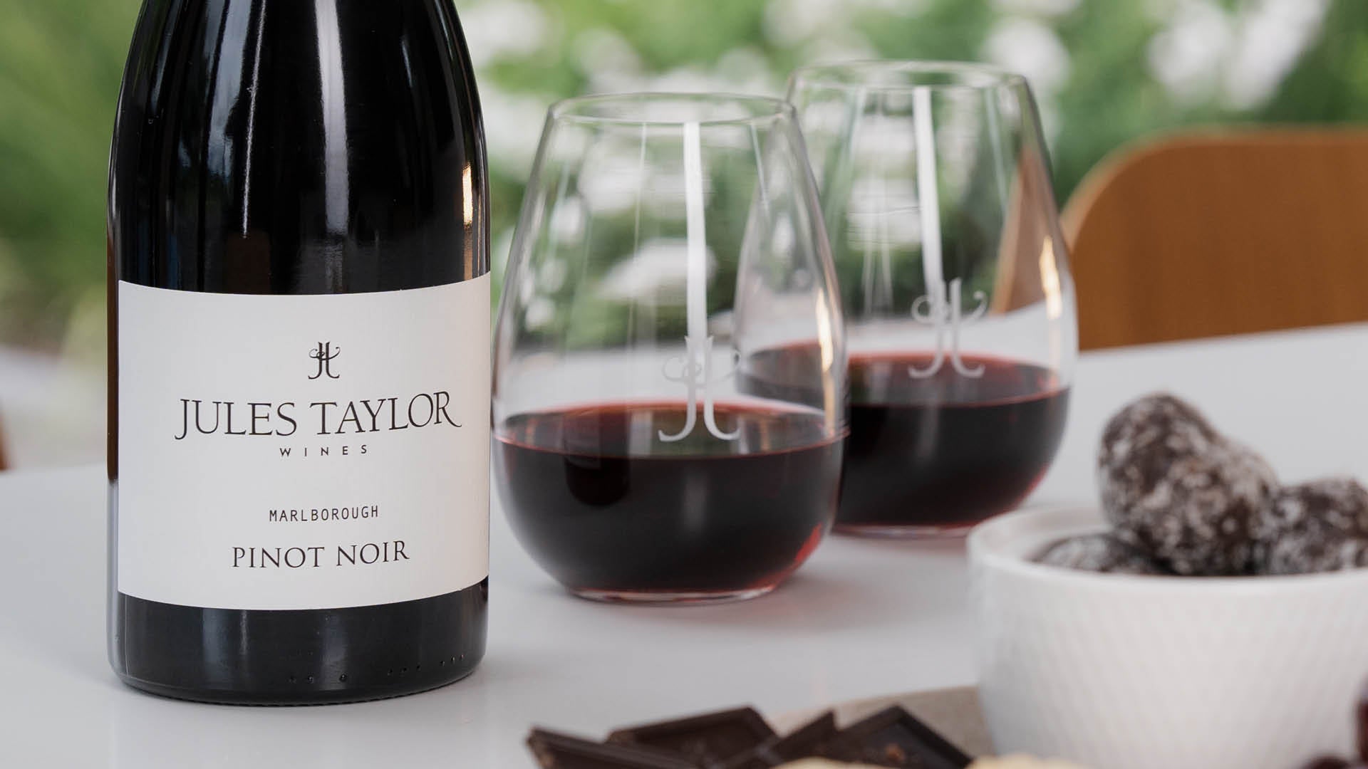 A bottle of Jules Taylor Wines Pinot Noir red wine stands on a white table next to two stemless glasses filled with wine, with chocolate snacks in the foreground and a blurred outdoor background.